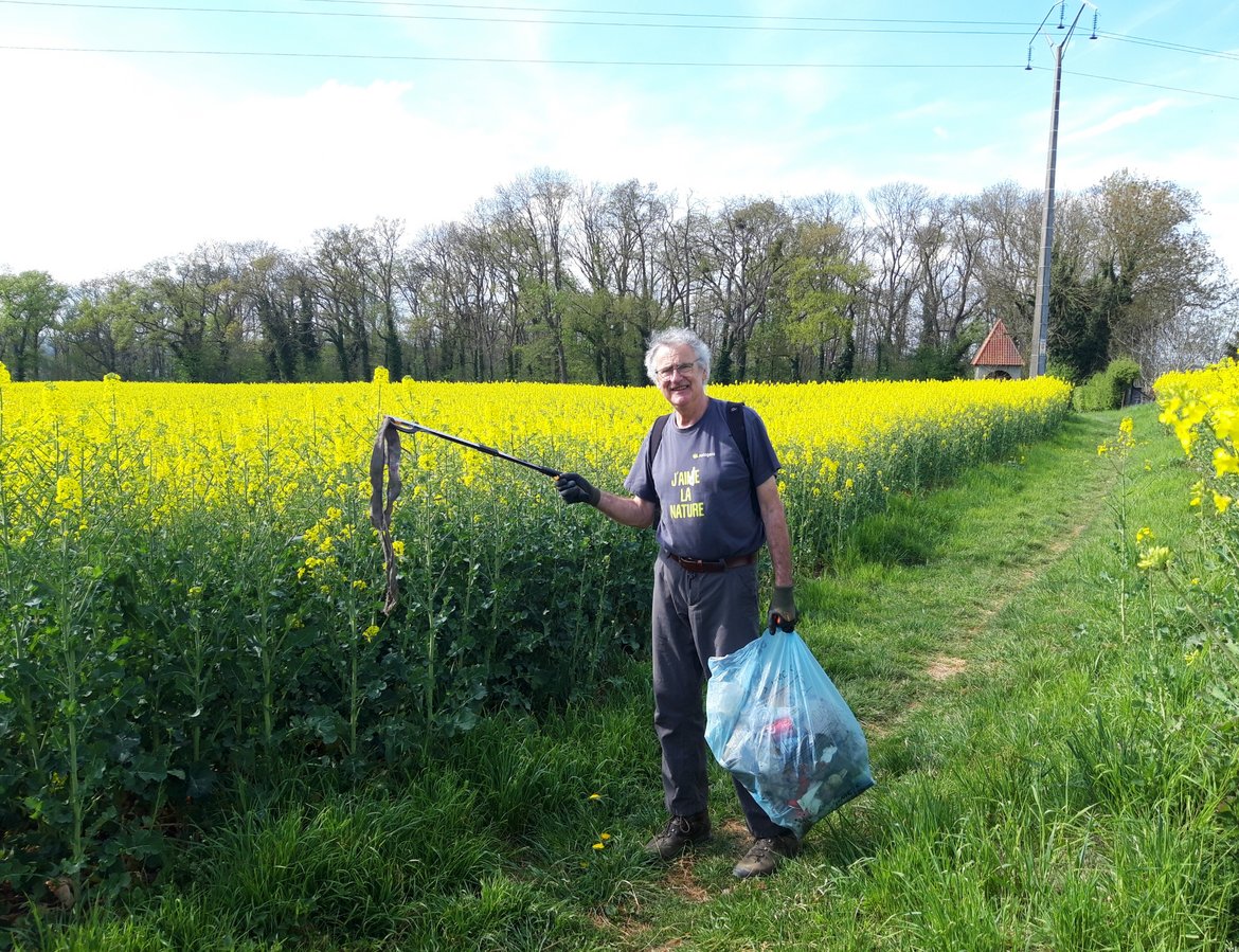 Sur un sentier de balade, au milieu d'un champs de fleurs jaunes, le colza, un volontaire ramasse un déchet